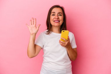 Elinde cep telefonu olan orta yaşlı beyaz kadın pembe arka planda izole edilmiş gülümseyen beş numarayı parmaklarıyla gösteriyor..