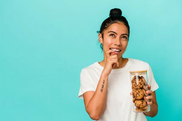 Young latin woman holding a cookies jar isolated on blue background relaxed thinking about something looking at a copy space.