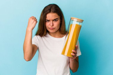 Mavi arka planda makarna kavanozu tutan genç beyaz kadın kameraya yumruk gösteriyor, agresif yüz ifadesi..
