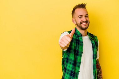 Sarı arka planda izole edilmiş genç dövmeli beyaz adam gülümsüyor ve başparmağını kaldırıyor.