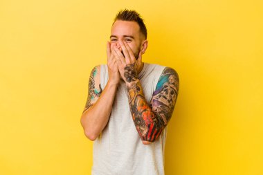 Sarı arka planda izole edilmiş genç dövmeli beyaz adam bir şeye gülüyor, elleri ağızda..