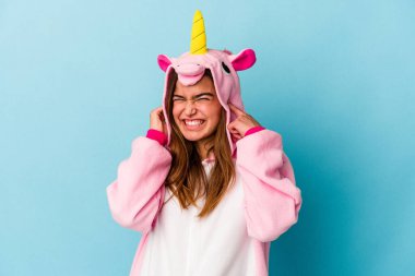 Tek boynuzlu pijama giyen beyaz bir kadın mavi arka planda izole edilmiş kulakları elleriyle kapatıyor..