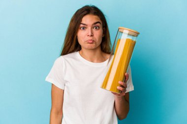 Mavi arka planda izole edilmiş makarna kavanozunu tutan beyaz bir kadın omuzlarını silker ve gözleri açık kalır..