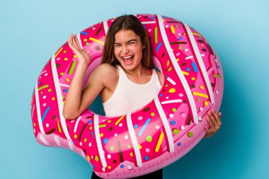 Mavi arka planda izole edilmiş, şişme donutlu genç beyaz kadın hoş bir sürpriz alıyor, heyecanlı ve el kaldırıyor..