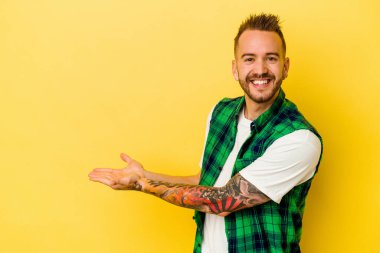 Sarı arka planda izole edilmiş genç dövmeli beyaz adam bir palmiye ağacının üzerinde bir kopya alanı tutuyor..