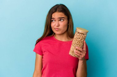 Beyaz arka planda izole edilmiş nohut kavanozu tutan genç beyaz adam şaşkın ve kararsız hissediyor..