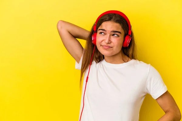 Young caucasian woman listening to music isolated on yellow background touching back of head, thinking and making a choice.