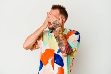 Beyaz arka planda izole edilmiş genç dövmeli beyaz adam elleriyle gözlerini kapatmaktan korkuyor..