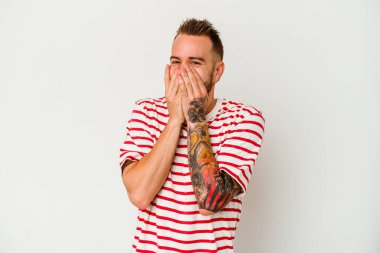 Beyaz arka planda izole edilmiş genç, dövmeli beyaz bir adam bir şeye gülüyor, elleriyle ağzını kapatıyor..