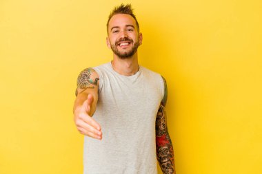 Sarı arka planda izole edilmiş genç dövmeli beyaz adam kameraya elini uzatarak selam veriyor..