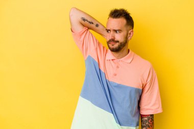 Genç dövmeli beyaz adam sarı arka planda izole edilmiş kafasının arkasına dokunuyor, düşünüyor ve bir seçim yapıyor..