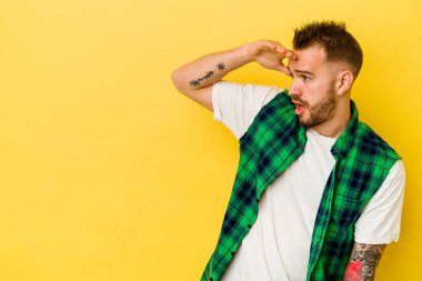 Genç dövmeli beyaz adam, sarı arka planda izole edilmiş. Uzağa bakıyor, elini alnına koyuyor..