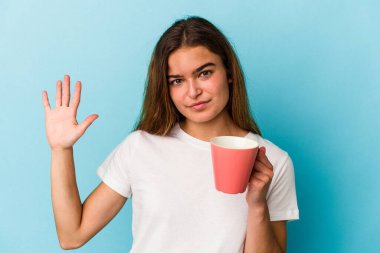 Kafkasyalı genç bir kadın mavi arka planda izole edilmiş bir kupa tutuyor. Beş numarayı parmaklarıyla gülümsüyor..