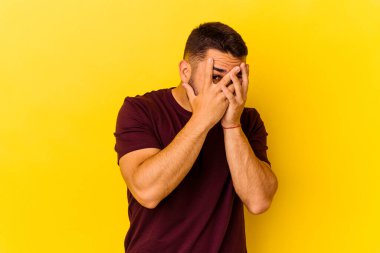 Sarı arka planda izole edilmiş genç beyaz adam parmaklarının arasından korkmuş ve gergin bir şekilde göz kırp..