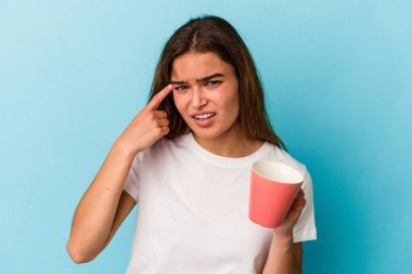 Kafkasyalı genç bir kadın mavi arka planda izole edilmiş bir kupa tutuyor işaret parmağıyla hayal kırıklığı jesti gösteriyor..