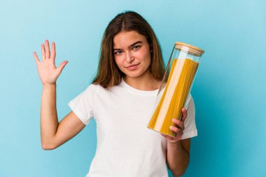 Mavi arka planda makarna kavanozu tutan beyaz bir kadın gülümseyerek beş numarayı parmaklarıyla gösteriyor..