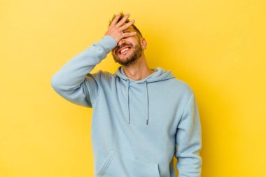 Sarı arka planda izole edilmiş genç dövmeli beyaz adam. Gözleri ellerle kaplıyor. Gülümsüyor ve bir sürpriz bekliyor..