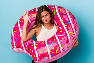 Şişme donutlu genç beyaz kadın mavi arka planda izole edilmiş. Beş numarayı parmaklarıyla gülümsüyor..