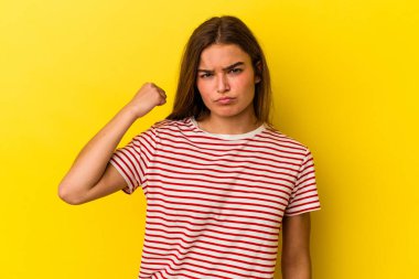 Sarı arka planda izole edilmiş genç beyaz kadın kameraya yumruğunu gösteriyor, agresif yüz ifadesi..