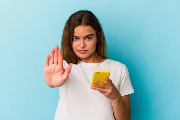 年轻的高加索女人拿着一部蓝色背景的手机站在那里 伸出了手 展示了停止的标志 阻止了你 — 图库照片