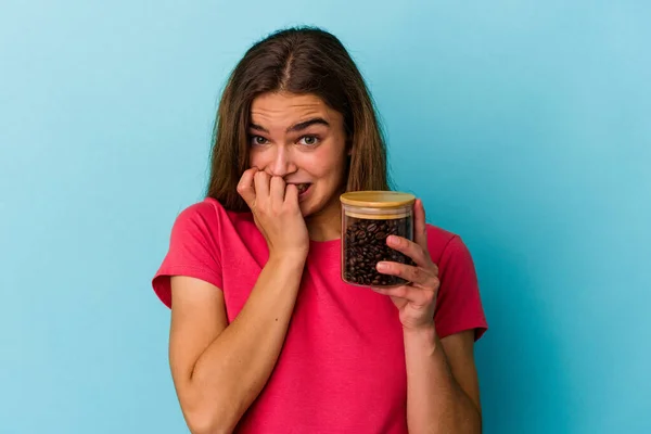 年轻的高加索女人拿着一个咖啡壶 用蓝色的背景隔离 咬着指甲 紧张而焦虑 — 图库照片
