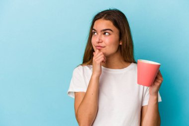 Kafkasyalı genç bir kadın mavi arka planda izole edilmiş bir kupa tutuyor şüpheli ve şüpheci bir ifadeyle yan bakıyor.
