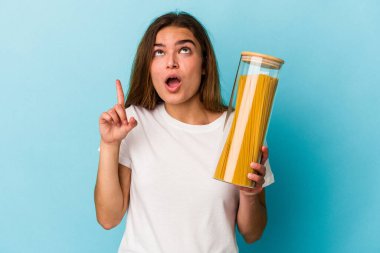 Elinde makarna kavanozu olan beyaz bir kadın mavi arka planda izole edilmiş ve ağzı açık bir şekilde üst tarafı işaret ediyor..