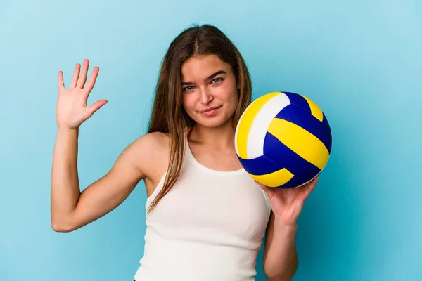 年轻的高加索女子在蓝色背景下打排球 开心地微笑着 用手指展示第五名 — 图库照片