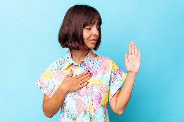 Genç melez kadın mavi arka planda izole edilmiş yemin ediyor, elini göğsüne koyuyor..