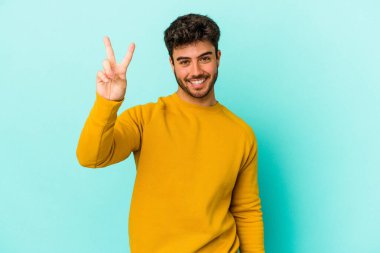 Kafkasyalı genç adam mavi arka planda izole edilmiş neşeli ve kaygısız parmaklarıyla barış sembolü gösteriyor..