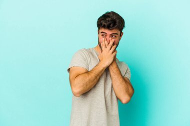 Mavi arka planda izole edilmiş genç beyaz bir adam bir şeye gülüyor, ağızlarını ellerle kapatıyor..
