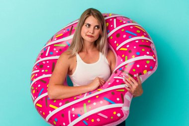 Mavi arka planda izole edilmiş, şişme donutu olan genç Rus kadın kafası karışmış ve kendinden emin değil..