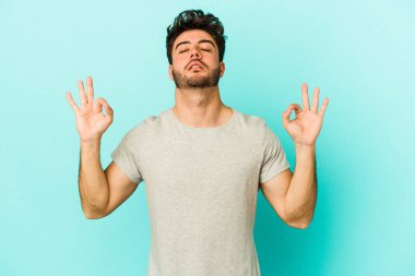 Mavi arka planda izole edilmiş genç beyaz adam yorucu bir günün ardından rahatlıyor. Yoga yapıyor..