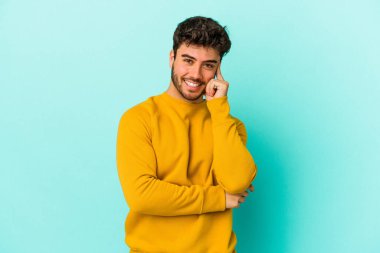 Mavi arka planda izole edilmiş genç beyaz adam gülümsüyor ve kendinden emin, elleriyle çenesine dokunuyor..