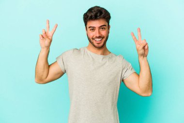Mavi arka planda izole edilmiş genç beyaz adam zafer işareti gösteriyor ve gülümsüyor..