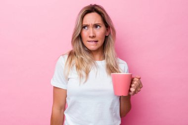 Pembe arka planda izole edilmiş pembe kupa tutan genç Avustralyalı kadın kafası karışmış ve kendinden emin değil..