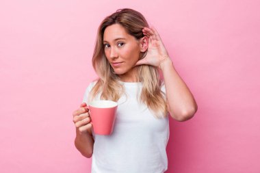 Pembe kupa tutan genç Avustralyalı bir kadın pembe arka planda izole edilmiş bir şekilde bir dedikoduyu dinlemeye çalışıyor..
