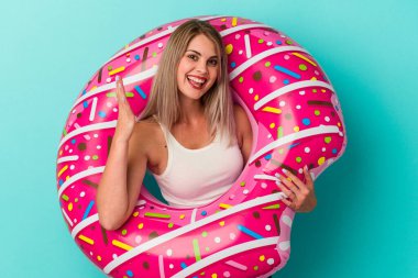 Mavi arka planda izole edilmiş şişme donutlu genç Rus kadın hoş bir sürpriz alıyor, heyecanlı ve el kaldırıyor..