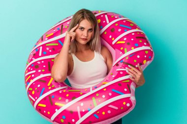 Şişme donutlu genç Rus kadın, mavi arka planda izole edilmiş, parmağıyla tapınağı işaret ediyor, düşünüyor, bir göreve odaklanıyor..