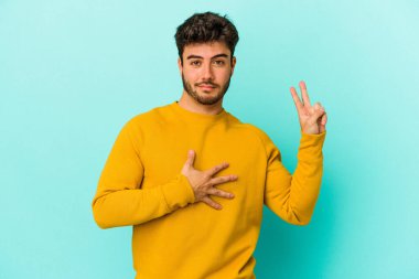 Kafkasyalı genç adam mavi arka planda izole edilmiş yemin ediyor, elini göğsüne koyuyor..