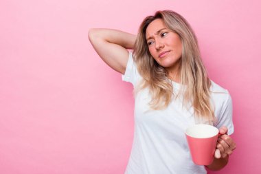 Genç Avustralyalı bir kadın pembe bir kupa tutuyor pembe arka planda izole edilmiş kafasının arkasına dokunuyor, düşünüyor ve bir seçim yapıyor..