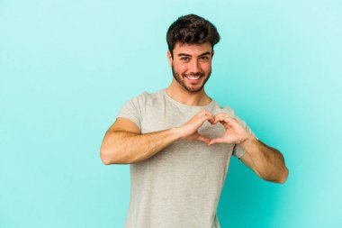 Mavi arka planda izole edilmiş genç beyaz adam gülümsüyor ve elleriyle kalp şeklini gösteriyor..