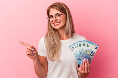 Genç Rus kadın pembe arka planda izole edilmiş banknotlar tutuyor gülümsüyor ve boş alanda bir şeyler gösteriyor..