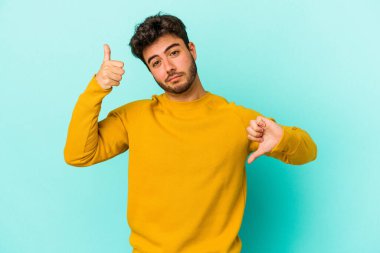 Kafkasyalı genç adam mavi arka planda izole edilmiş, baş parmağını kaldırıyor, zor seçim konsepti.