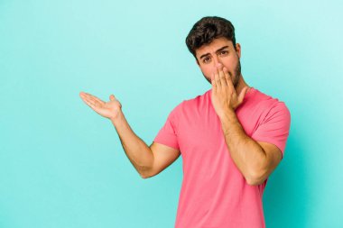 Mavi arka planda izole edilmiş genç beyaz adam, avuç içi fotokopi alanını elinde tutmaktan etkilenmiş..