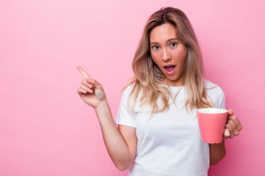 Pembe arka planda izole edilmiş pembe kupa tutan genç Avustralyalı kadın yan tarafı işaret ediyor.