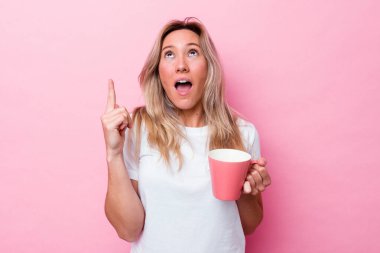 Genç Avustralyalı bir kadın pembe bir kupa tutuyor. Pembe arka planda izole edilmiş, ağzı açık, üst tarafı işaret ediyor..