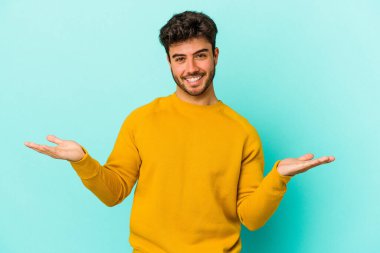 Mavi arka planda izole genç beyaz adam kollarıyla ölçek yapar, mutlu ve kendinden emin hisseder..