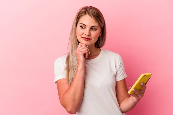 年轻的俄罗斯女人拿着一部粉色背景的手机 侧着眼睛 带着怀疑和怀疑的表情 — 图库照片