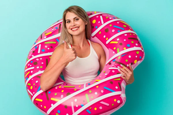 Şişme donutlu genç Rus kadın mavi arka planda izole edilmiş gülümsüyor ve başparmağını kaldırıyor.